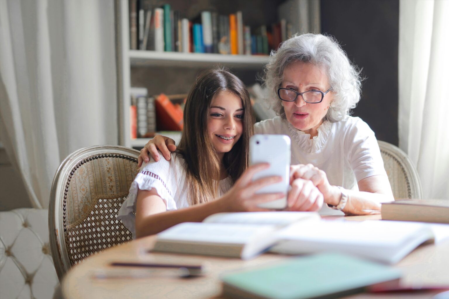 A grandmother and granddaughter sitting at a table with books and letterwriting materials while looking at a phone together.