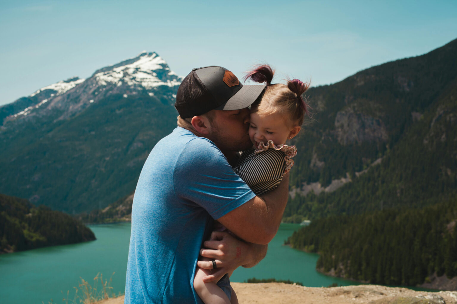 A father hugging and kissing his daughter in a mountain landscape with a lake.
