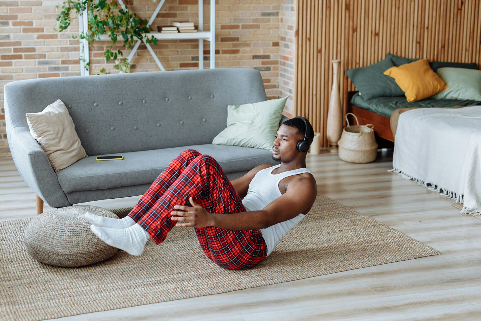 A man exercising on a rug by a couch while wearing plaid pajama pants, a tank top, and headphones.