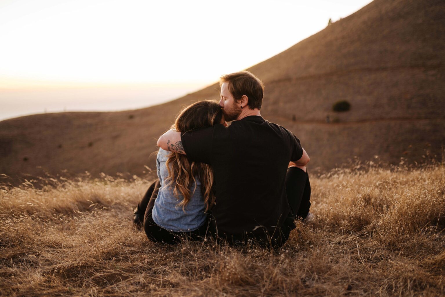 A man kissing a woman’s forehead while sitting together on a grassy hill at sunset.