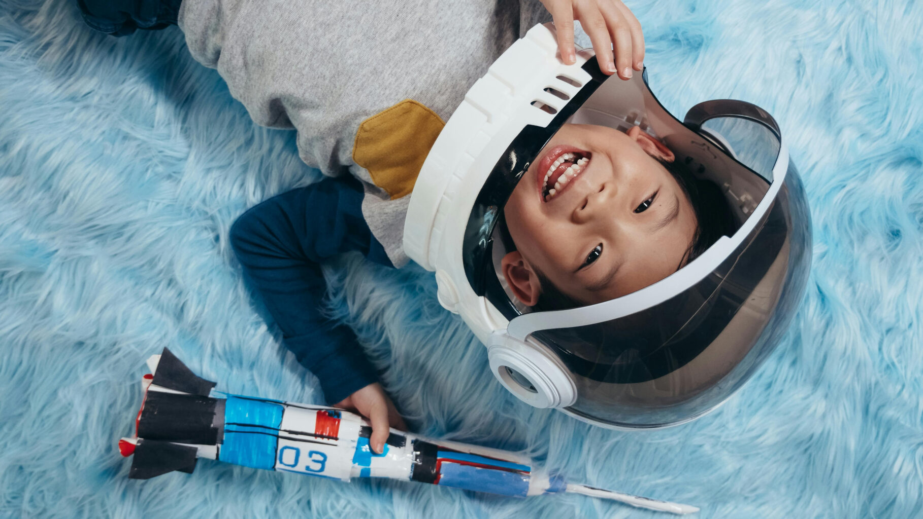 A boy in an astronaut helmet holding a rocket while lying on a blue fur blanket.