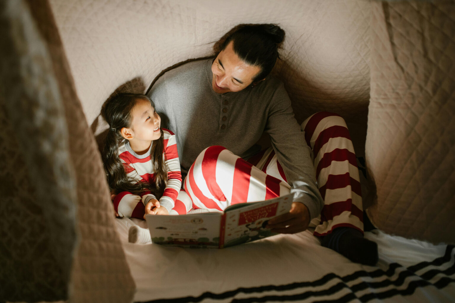 A father and daughter wearing striped pajamas and reading a book under a blanket