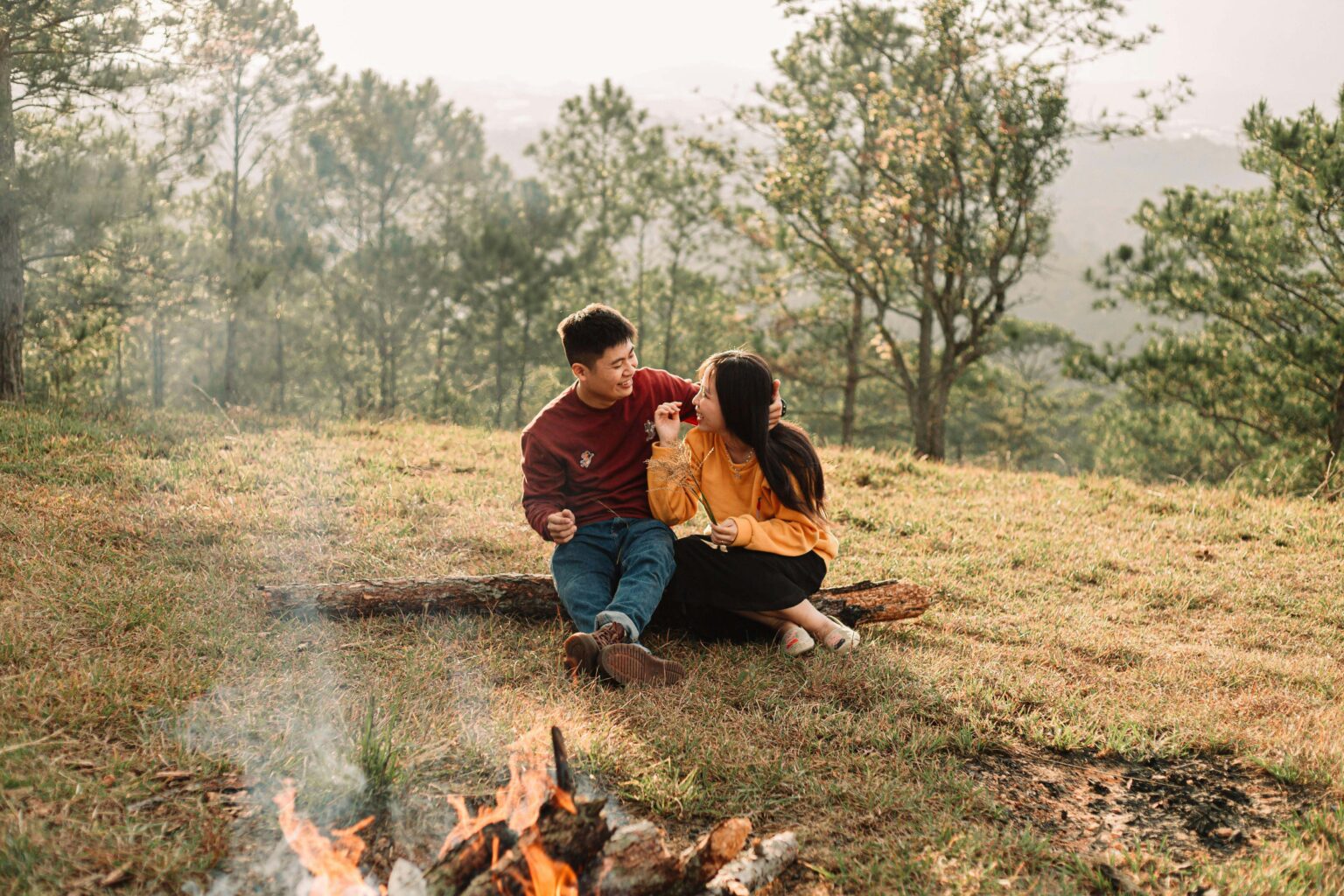 A couple sitting on a log by a fire in the woods.