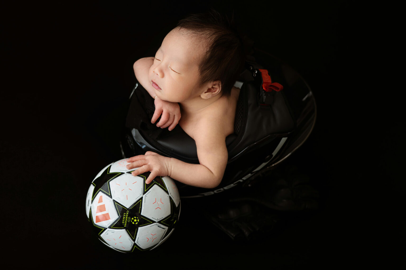 A newborn sleeping while holding a soccer ball with one hand.