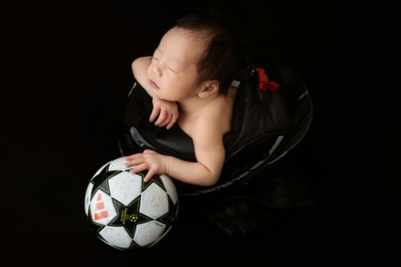 A newborn sleeping while holding a soccer ball with one hand.