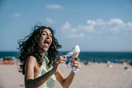 A woman holding an ice cream cone on the beach and laughing.