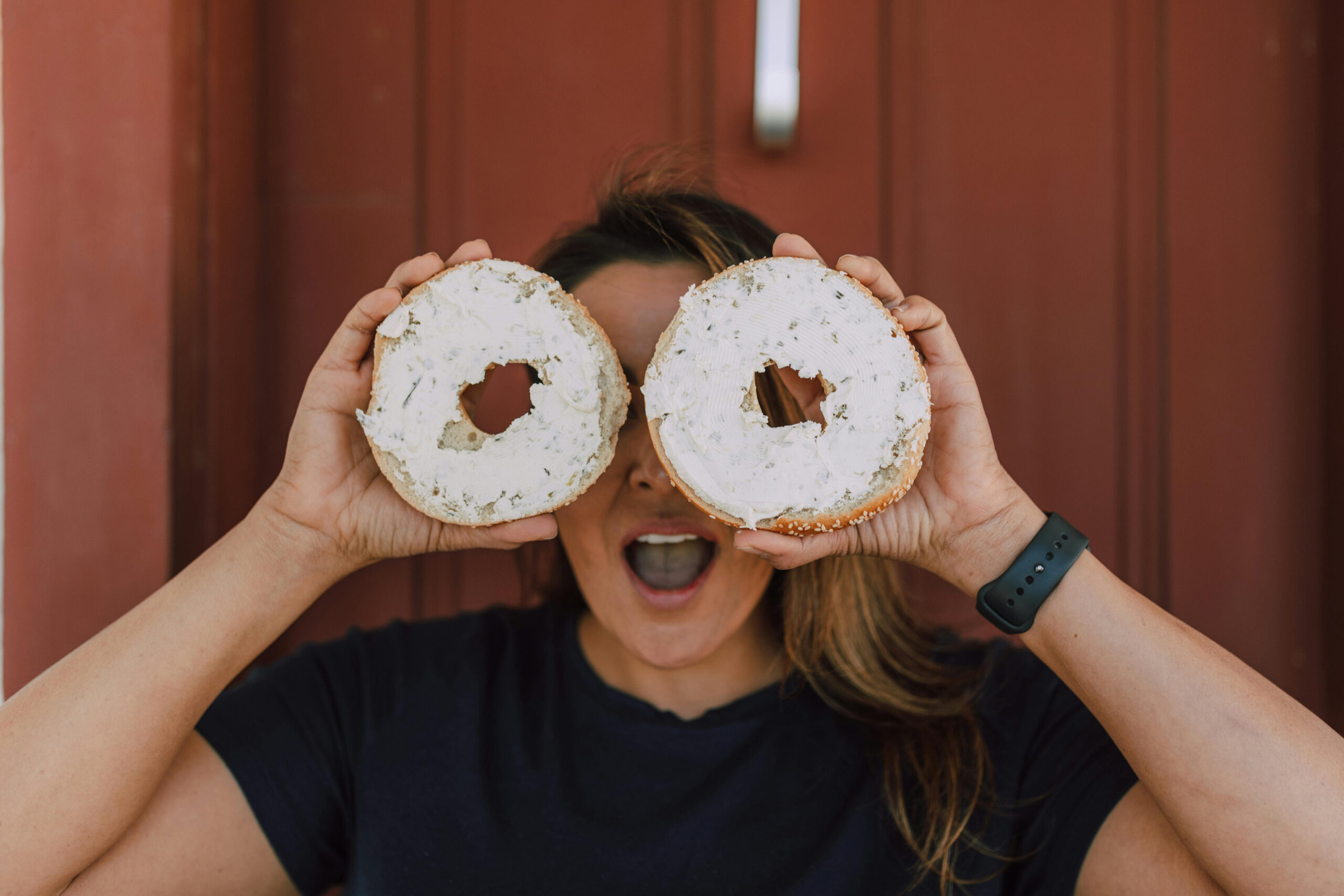 A woman holding bagels with cream cheese over her eyes.